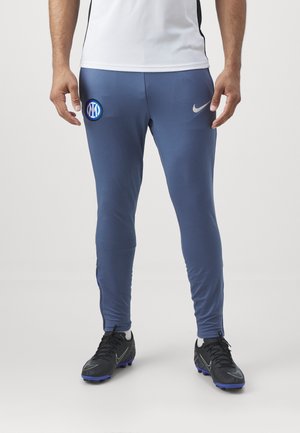 Partie inférieure du corps d'une personne portant un pantalon d'entraînement bleu de l'Inter Milan avec le logo Nike et des crampons de football noirs sur un fond blanc.