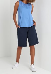 Blaues ärmelloses Top mit strukturiertem Ausschnitt, kombiniert mit dunkelblauen knielangen Shorts und weißen Sneakers, vor einem hellen Hintergrund.