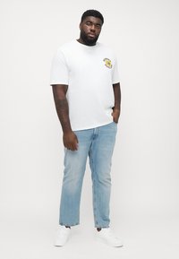 White short-sleeve T-shirt featuring a yellow graphic printed on the chest, paired with light blue jeans and white sneakers.