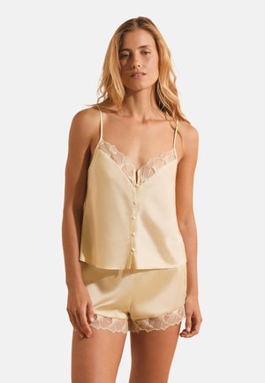 Giovane donna con canottiera in raso beige con bordi in pizzo e shorts coordinati su sfondo bianco semplice.