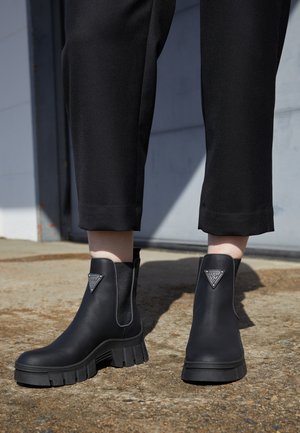 Personne portant des bottines noires à semelle épaisse avec le logo Guess, associées à un pantalon noir raccourci, debout sur un sol texturé en plein air.