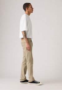 Camiseta blanca de manga corta, pantalones beige ajustados, zapatillas negras de corte bajo con suelas blancas. Presenta un tatuaje en el brazo izquierdo.