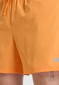 Oranssit urheilushortsit, joissa on joustava vyötärö ja kiristysnyöri. Shortsien pinta on sileä ja sivussa on heijastava logo.