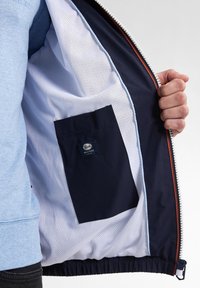 Marineblauwe jasinterieur met een gaasvoering en een donkerblauwe pocket, voorzien van een label. De buitenkant heeft een rits met een contrasterende oranje accent.