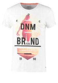 Maglietta bianca con una scena di spiaggia al tramonto, silhouette di una donna e il testo "DNM BRND co. SUMMER BREEZE 1998" sulla parte anteriore.