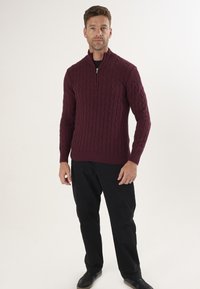 Bordowy sweter w warkoczowy splot z kołnierzem na pół zamek, ściągacze na mankietach i u dołu, w połączeniu z czarnymi spodniami i ciemnymi sneakersami.