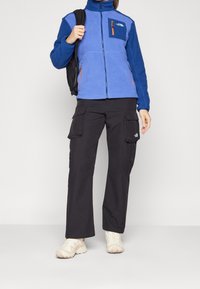 Niebieska polarowa kurtka z zamkiem błyskawicznym na piersi, połączona z czarnymi spodniami cargo z wieloma kieszeniami. Beżowe buty sportowe dopełniają cały zestaw.