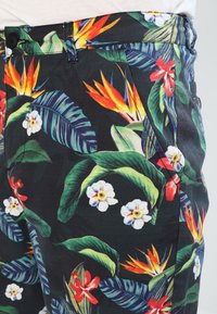 Pantaloni da uomo realizzati in tessuto con uno sfondo nero e un vivace motivo floreale tropicale, che include foglie, orchidee e uccelli del paradiso.
