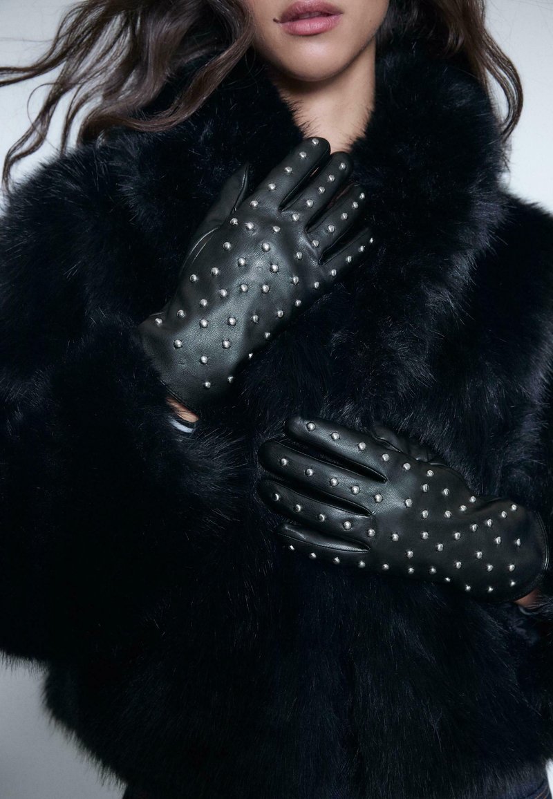 Schwarze Lederhandschuhe mit silbernen Nieten, die ein elegantes Design aufweisen. Getragen mit einem strukturierten schwarzen Pelzmantel, der einen gewagten Look unterstreicht.