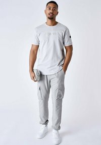 Camiseta de manga corta gris claro con logo en relieve, combinada con pantalones cargo gris claro que tienen bolsillos y puños. Los accesorios incluyen un estuche a juego.