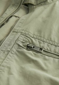Veste vert clair dotée d'une texture de tissu lisse, d'une poche zippée bien marquée et de coutures détaillées le long des coutures et des zips.