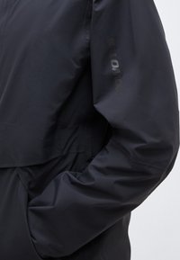 Manche de veste noire imperméable avec une main partiellement dans la poche latérale, affichant un logo vertical discret "SALOMON" sur le haut du bras.