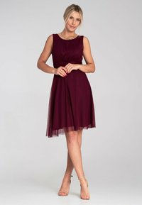 Burgunderfarbenes ärmelloses Kleid mit tailliertem Oberteil, geraffter Taille und einem Tüllrock. Getragen mit nudefarbenen, hohen Sandalen und einer schlichten Halskette.