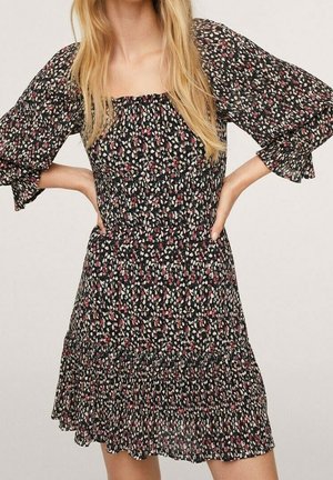 Femme portant une robe noire à motifs floraux avec de petites fleurs roses et blanches, décolleté carré et manches bouffantes trois-quarts.
