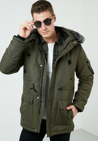 Parka verde con capucha forrada de pelo, bolsillos delanteros con broche, cremalleras verticales y cordones ajustables en la cintura y los puños.