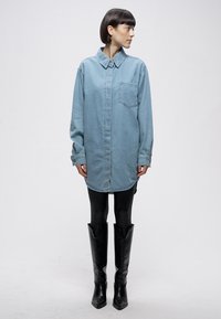 Abito camicia in denim azzurro chiaro con chiusura a bottoni, maniche lunghe, taschino sul petto e orlo arrotondato, abbinato a stivali neri lucidi al ginocchio.