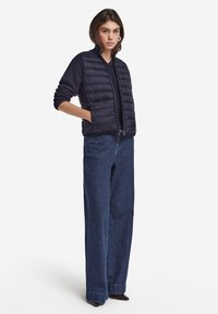 Marinblå pufferväst med horisontell sömnad, dragkedja fram och sidofickor, tillsammans med högmidjade blå denimbyxor. Slät tygtextur.