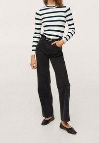Schwarze hochgeschnittene Jeans mit geraden Beinen, kombiniert mit einem langärmeligen Oberteil in Weiß und Marineblau mit Streifen. Flache schwarze Schuhe mit einer Schleifenakzent.