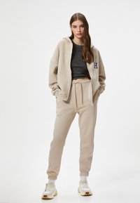Beige zip-up hoodie med store frontlommer og broderet logo, parret med matchende joggers med elastiske manchetter, båret med sneakers.