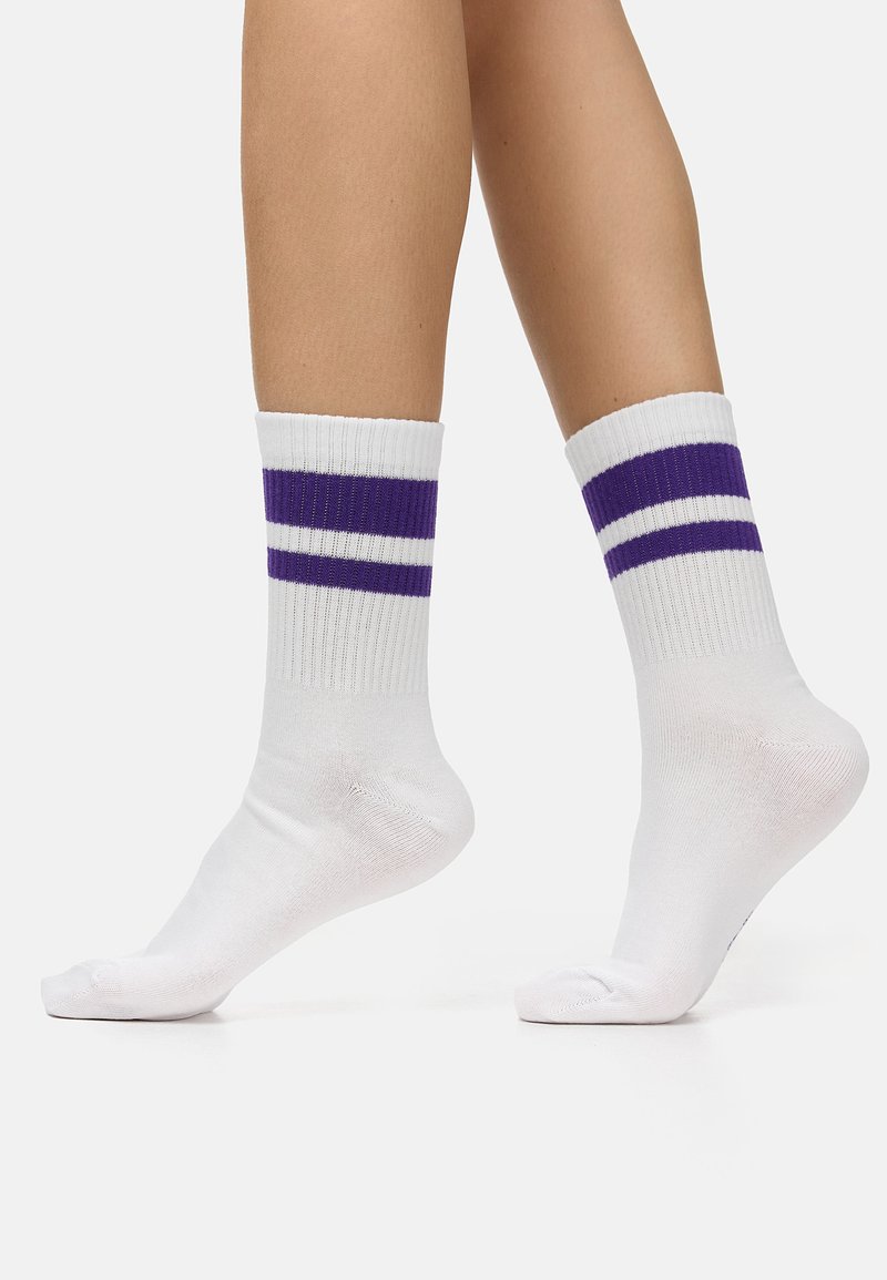 Nogi w białych skarpetkach typu crew z dwoma poziomymi fioletowymi paskami u góry, stojące na białej, gładkiej powierzchni.