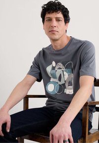 Pilkas trumpas rankovės t-shirt su abstrakčiu dizainu, kuriame yra apskritimų ir ovalių formų santūriomis spalvomis. Lygus audinys, laisvas siluetas.