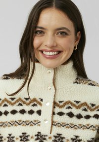 Pull en polaire crème avec des motifs géométriques marron et noir. Présente un col haut et des boutons-pression argentés sur le devant.