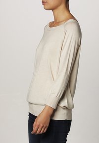 Beige pletený svetr s dlouhými rukávy, širokým výstřihem ve tvaru loku a ribbenými manžetami. Uvolněný střih s hladkou texturou.