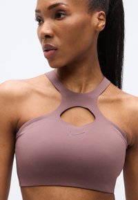 Αθλητικό σουτιέν σε ανοιχτό μωβ χρώμα από ελαστικό ύφασμα. Διαθέτει σχέδιο με ανοίγματα και στυλ racerback. Μικρό λογότυπο Nike στο μπροστινό μέρος.