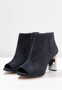 Botas de tobillo de ante azul marino, abiertas en la punta, con un tacón bloque de acrílico plateado y cierre de cremallera lateral. Textura suave y diseño elegante.