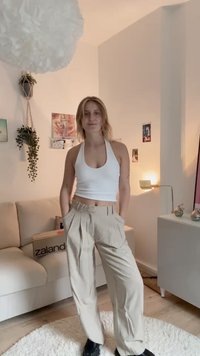 Weißes Hals-top kombiniert mit beigen, plissierten Hosen. Der Raum zeichnet sich durch helle Farben, eine Pflanze und dekorative Wandkunst aus. Glatte Texturen und klare Linien.