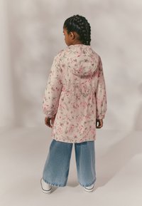 Veste imperméable rose à motif floral avec capuche, poignets élastiques et taille cintrée. Associée à un jean large en denim et des chaussures blanches.