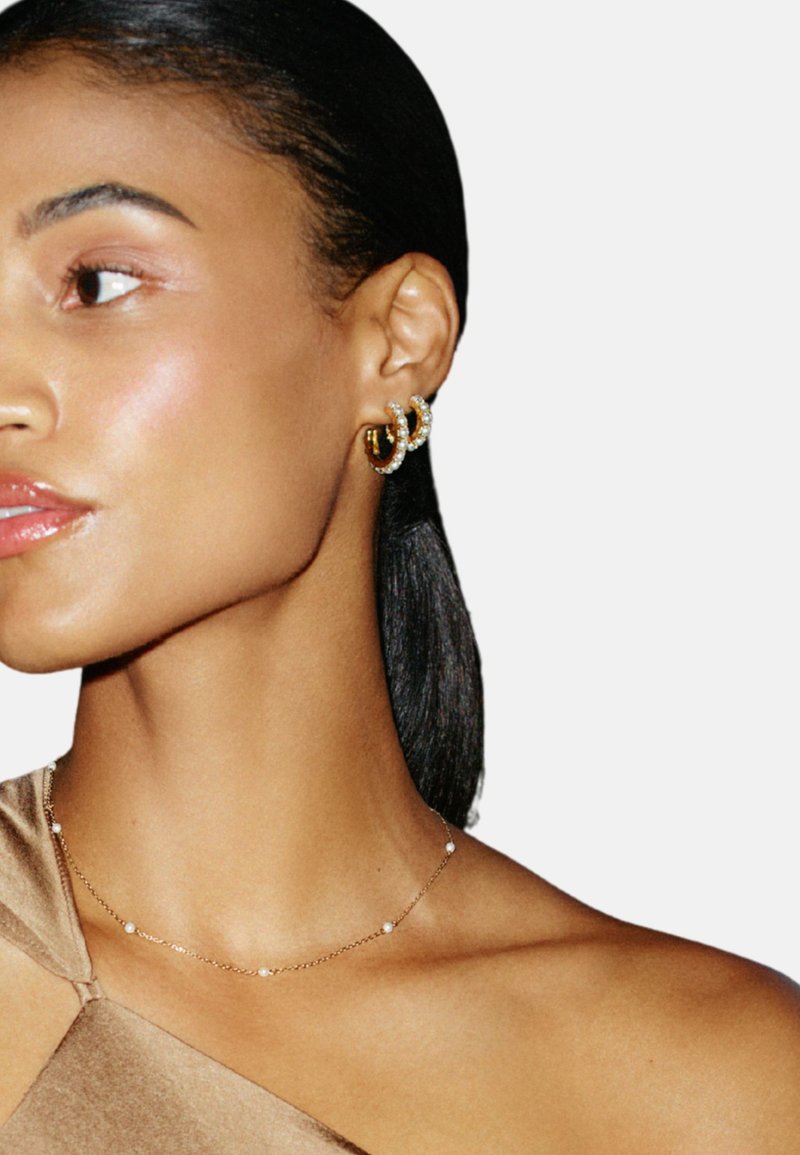 Guldörhängen i ringform med kristalldetaljer och en texturerad design. Ett tunt guldkedjehalsband med små vita pärlor är också synligt.