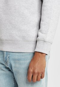 Felpa grigia realizzata in tessuto morbido con polsini e orlo a coste, abbinata a jeans in denim di un blu chiaro. Primo piano di una mano appoggiata sui pantaloni.