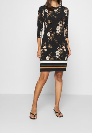 Femme portant une robe noire mi-longue avec des motifs floraux beiges et blancs et des rayures horizontales à l’ourlet, associée à des sandales noires.