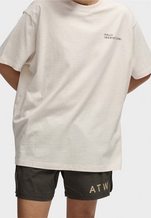 Helle beige, oversized T-Shirt aus weichem Stoff mit Rundhalsausschnitt und kleinem schwarzen Druck. Kombiniert mit dunkelgrünen Shorts mit goldfarbenem Logo.