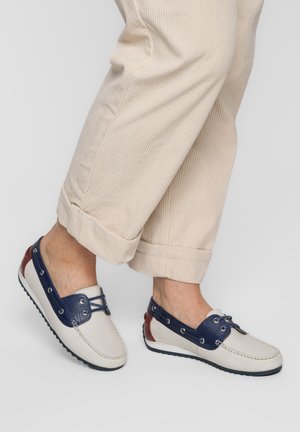 Personne portant un pantalon en velours côtelé beige et des mocassins blancs avec des accents bleu marine et rouges, debout devant un fond clair uni.