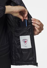 Interno di una giacca nera isolata con una tasca sicura che contiene uno smartphone blu. Etichetta bianca con logo rosso visibile. Dettaglio con zip incluso.