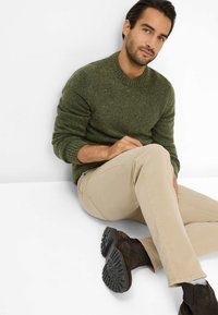 Maglione verde lavorato a maglia con scollo rotondo, pantaloni beige e stivaletti neri alla caviglia. Modella seduta su uno sfondo bianco.