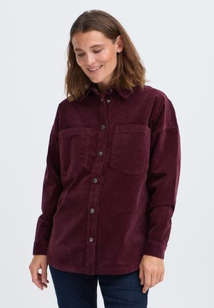 Femme aux cheveux bruns mi-longs portant une chemise boutonnée bordeaux foncé à manches longues et un pantalon sombre, souriante et regardant vers le bas.