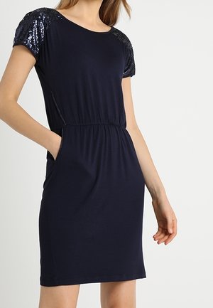 Woman wearing a knee-length navy dress with sequin short sleeves and side pockets, standing with one hand in a pocket.