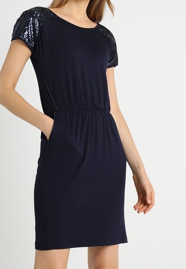 Femme portant une robe marine mi-longue avec des manches courtes à sequins et des poches latérales, debout avec une main dans une poche.