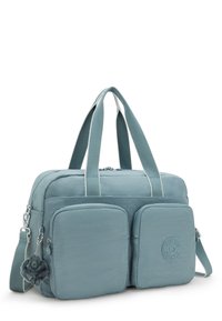 Blue fabric bag with dual front pockets, silver hardware, detachable furry keychain, dual handles, and adjustable shoulder strap.