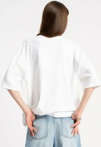 Kobieta z długimi brązowymi włosami, ubrana w luźną białą koszulę z krótkim rękawem i jasnoniebieskie dżinsy, z rękami wsuniętymi w tylne kieszenie.