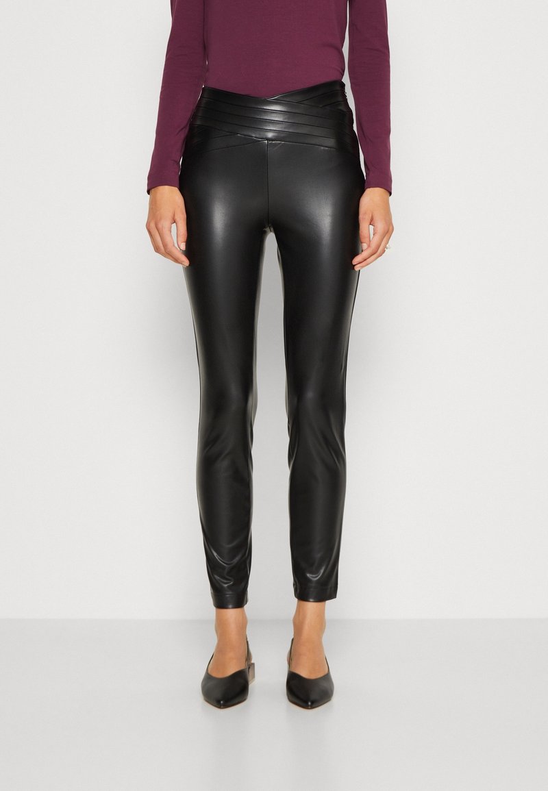 Personne portant un pantalon noir en cuir synthétique taille haute avec des détails de panneaux horizontaux et des talons noirs pointus, debout devant un fond blanc.