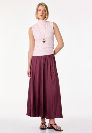 Woman standing wearing sleeveless pale pink high-neck top, long maroon skirt, pendant necklace, and black flat sandals.