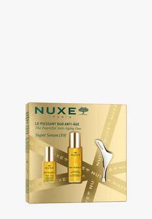 Das NUXE Anti-Aging-Duo in einer goldenen Box enthält zwei Amber-Serumfläschchen mit elegantem Design und ein silbernes Applikatorwerkzeug. Der Text enthält Produktdetails.
