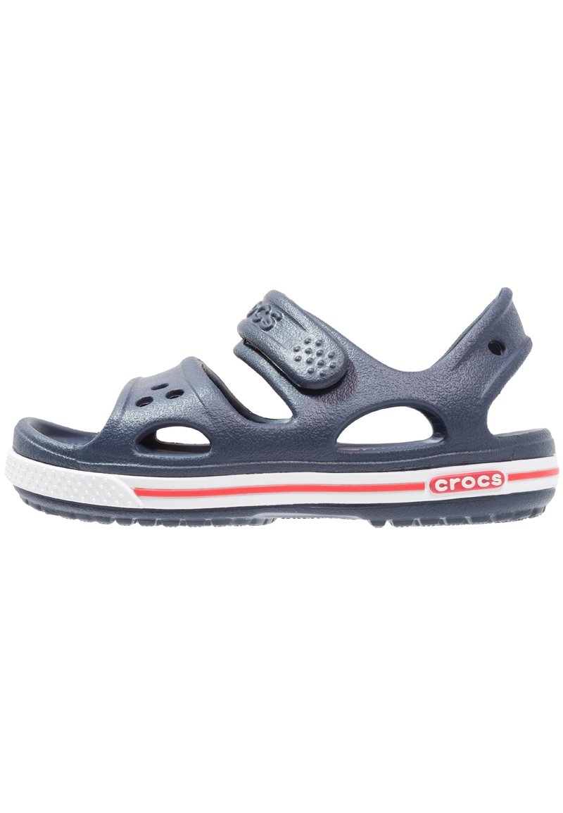 Tummansiniset Crocs-sandaalit säädettävällä hihnalla, avoimella muotoilulla, ilmanrei'illä, valkoisella välipohjalla ja punaisilla korostusviivoilla ulkopohjassa.