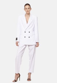 Kobieta stoi w białym podwójnym żakiecie i dopasowanych spodniach, w połączeniu z czarnymi sandałami na wysokim obcasie, zwrócona przodem.