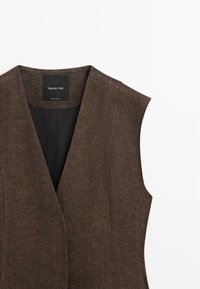 Donkerbruine mouwloze vest met textuur, diepe V-hals en een label met "Massimo Dutti" binnenin de kraag.