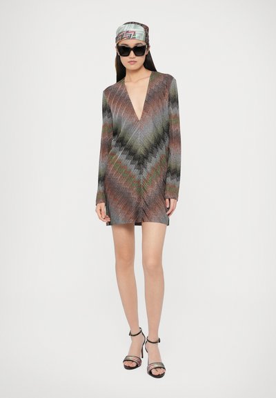 Pletené šaty s dlhými rukávmi, hlbokým výstrihom do V, cik-cak vzor v odtieňoch sivej, zelenej a červenej. Doplnky zahŕňajú čierne slnečné okuliare a viacfarebný šál na hlavu.